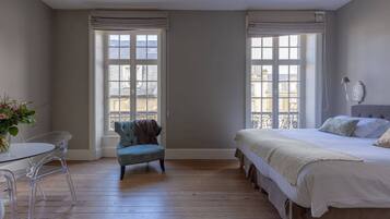 A bedroom with a bed, a chair, a table, and two windows with curtains.