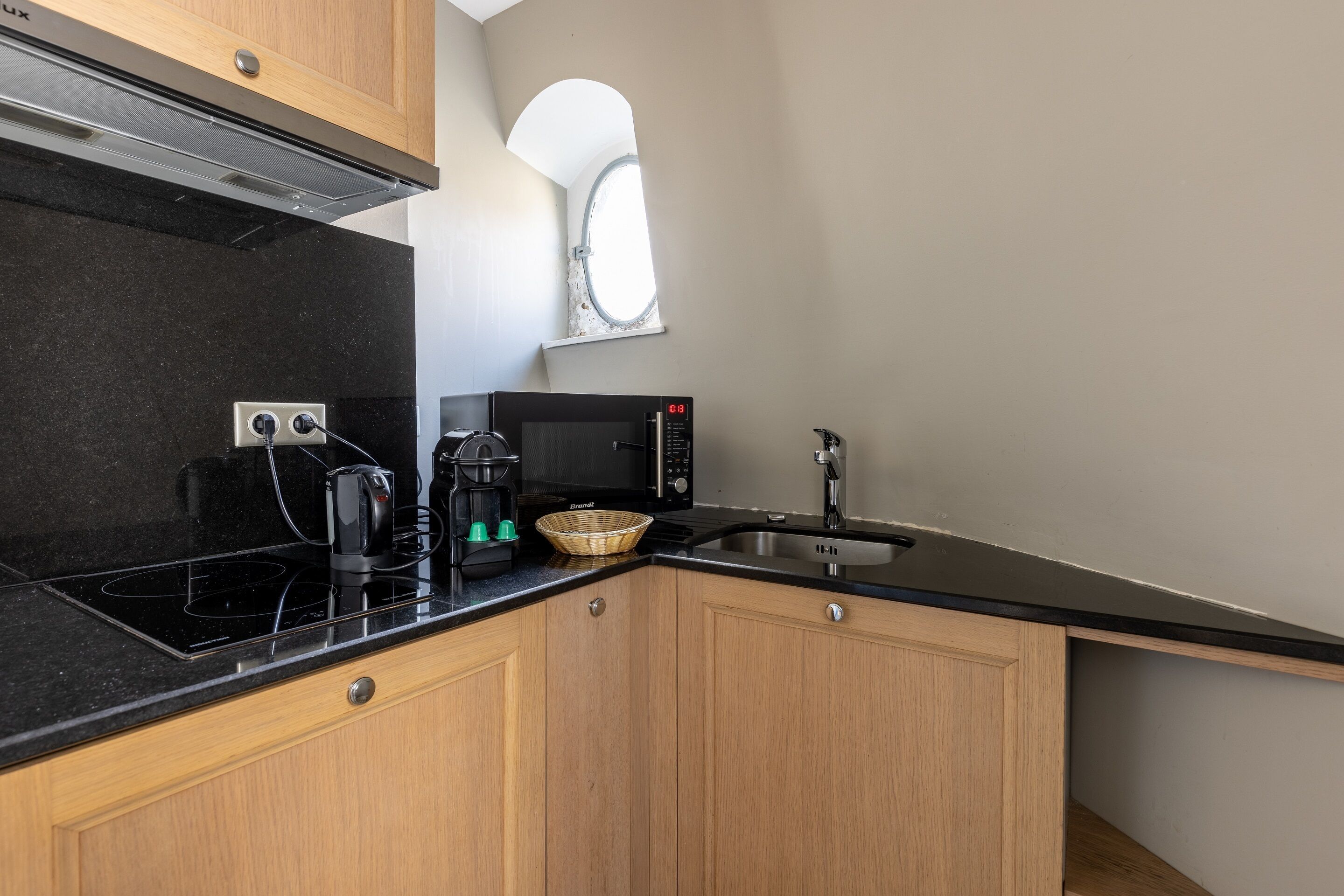 Una cocina compacta con cubierta negra, armarios de madera, microondas, cafetera y ventana sobre el fregadero.