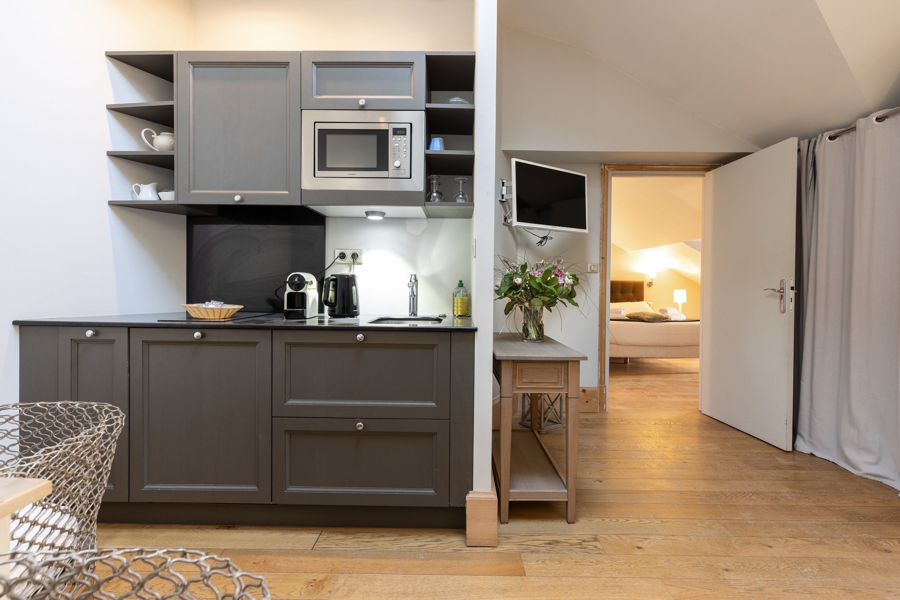Cocina compacta con gabinetes grises, microondas y cafetera. Hay una pequeña mesa con una planta y una silla. El cuarto tiene piso de madera y una puerta que da a otra área.