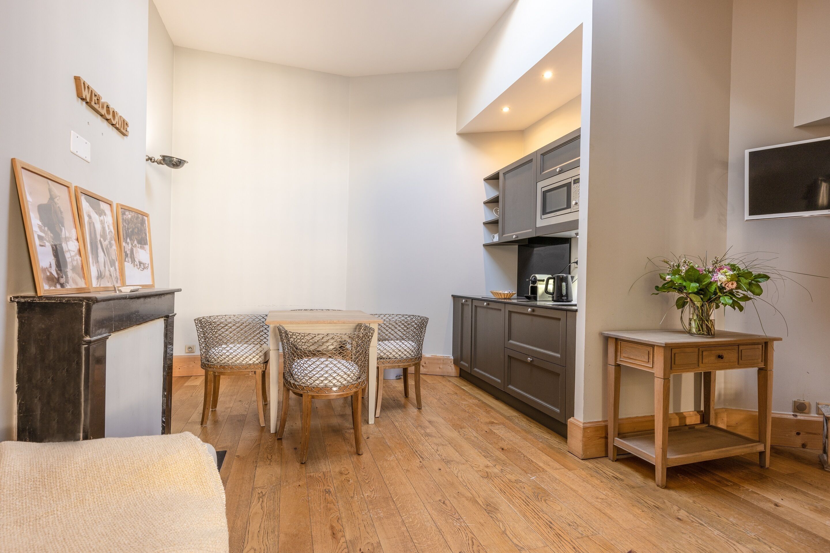 A compact kitchen with a microwave, a small table with chairs, a wooden side table with a vase, and a 'WELCOME' sign on the wall.