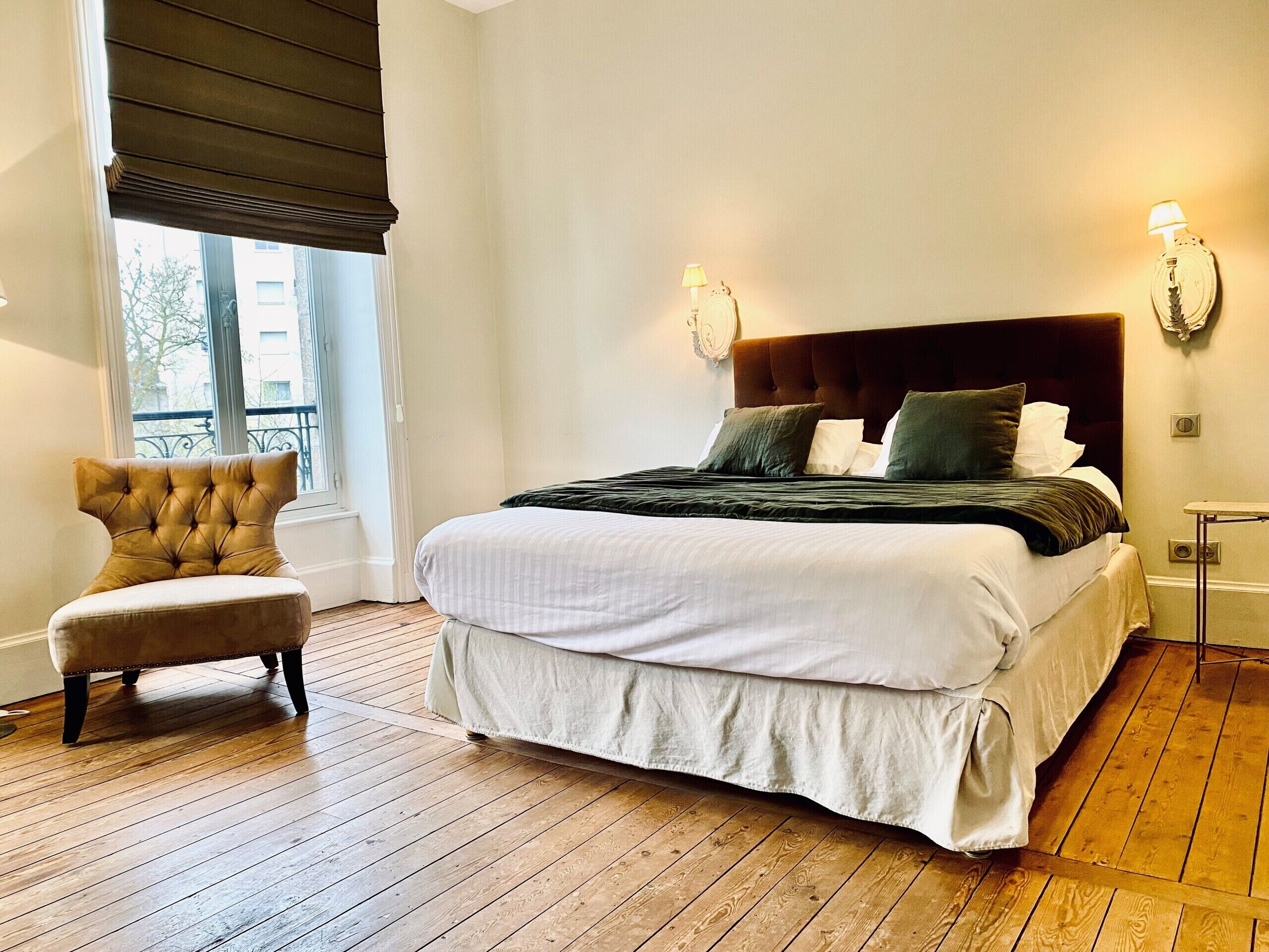 A neatly made bed with a headboard, a chair, and a window with blinds.