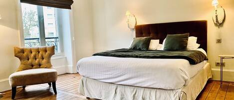 A neatly made bed with a headboard, a chair, and a window with blinds.