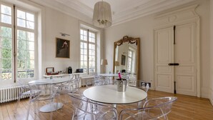 Une pièce spacieuse dotée de grandes fenêtres, d’un lustre et d’un coin repas avec des chaises transparentes et une table blanche.