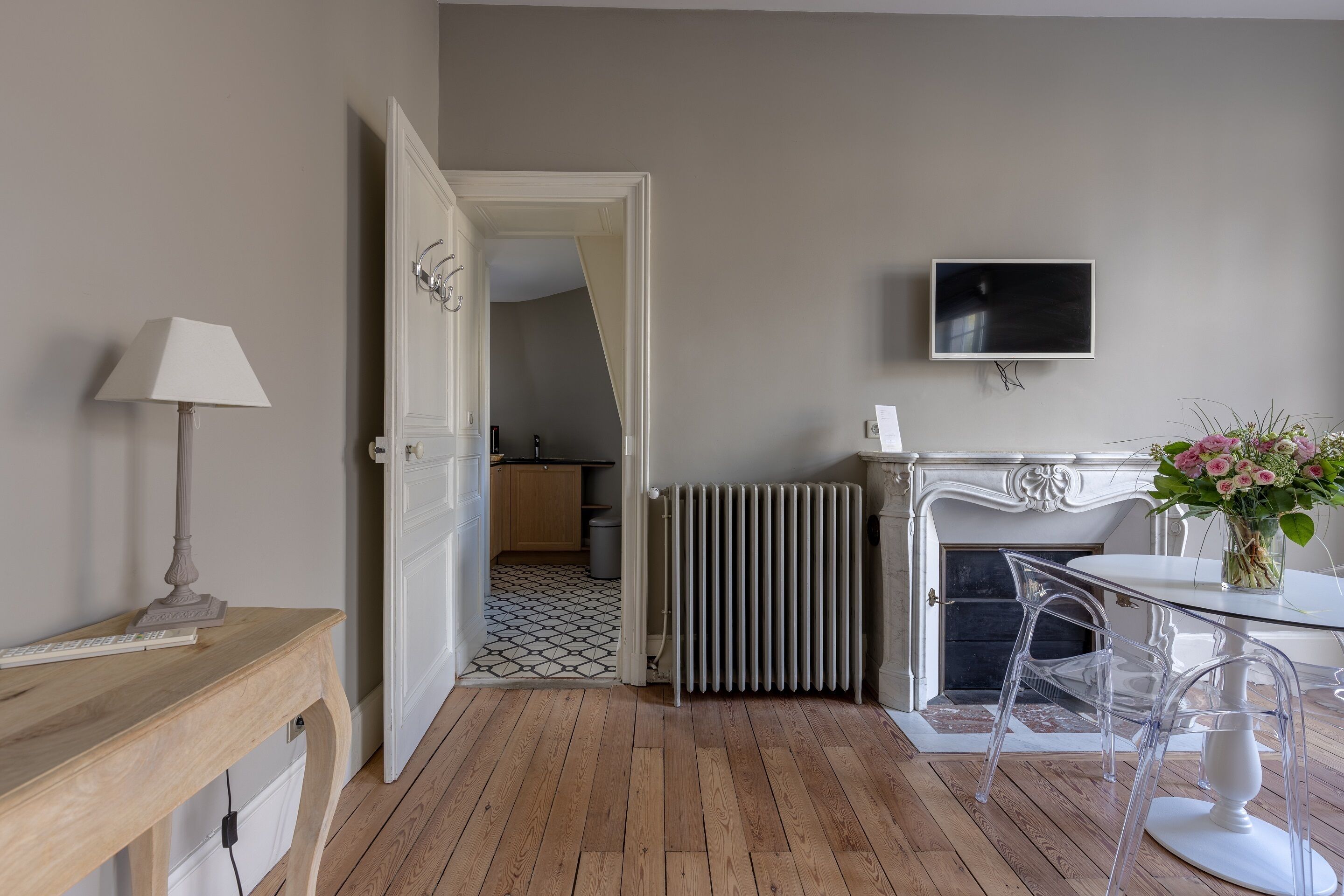 Una habitación con suelo de madera, un radiador blanco, una mesa de vidrio transparente con sillas, una lámpara blanca y un televisor en la pared.