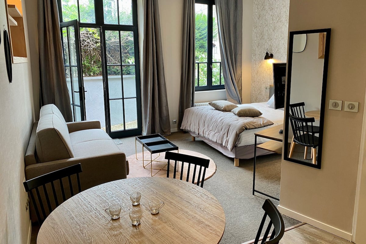 A modern room with a bed, a sofa, a dining table with chairs, and a large window with curtains.