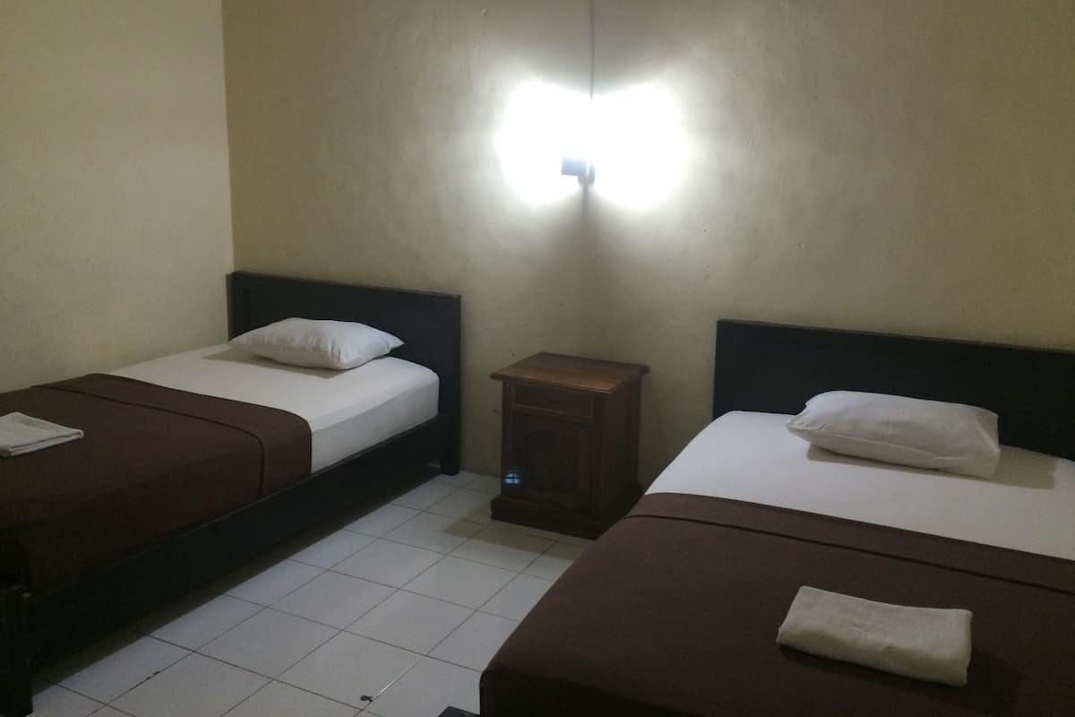 A hotel room with two single beds, each with a pillow and a folded towel. There is a small wooden nightstand between the beds.