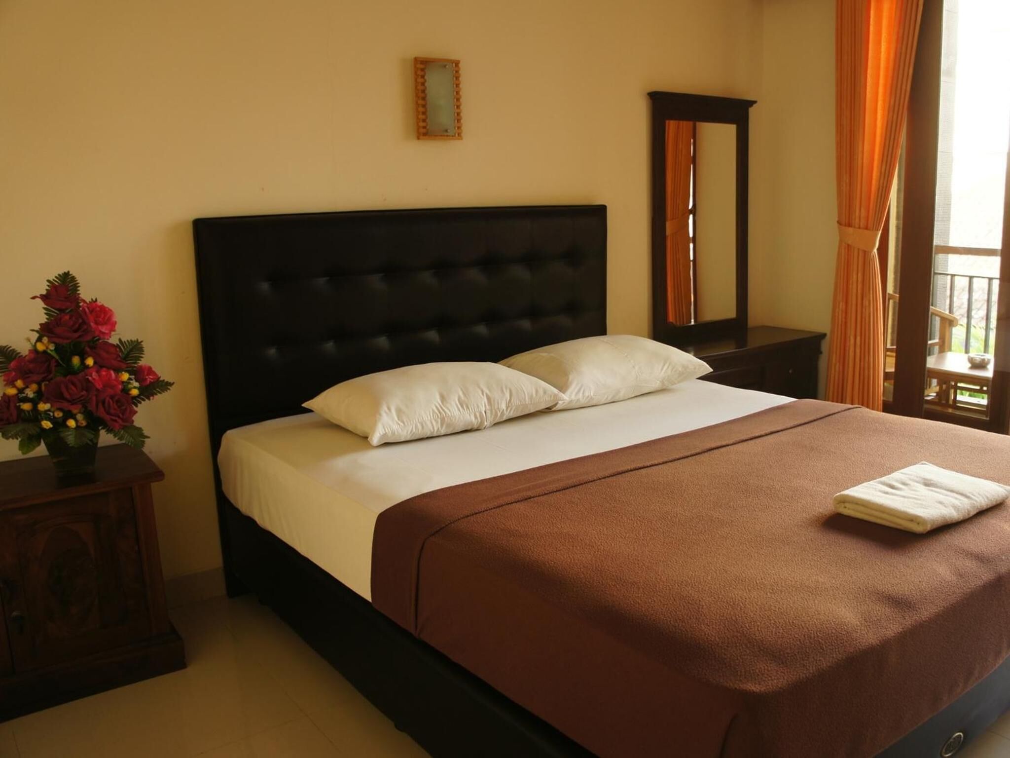 A neatly made bed with a dark headboard, white pillows, and a brown bedspread. There is a wooden nightstand with a vase of flowers to the left.