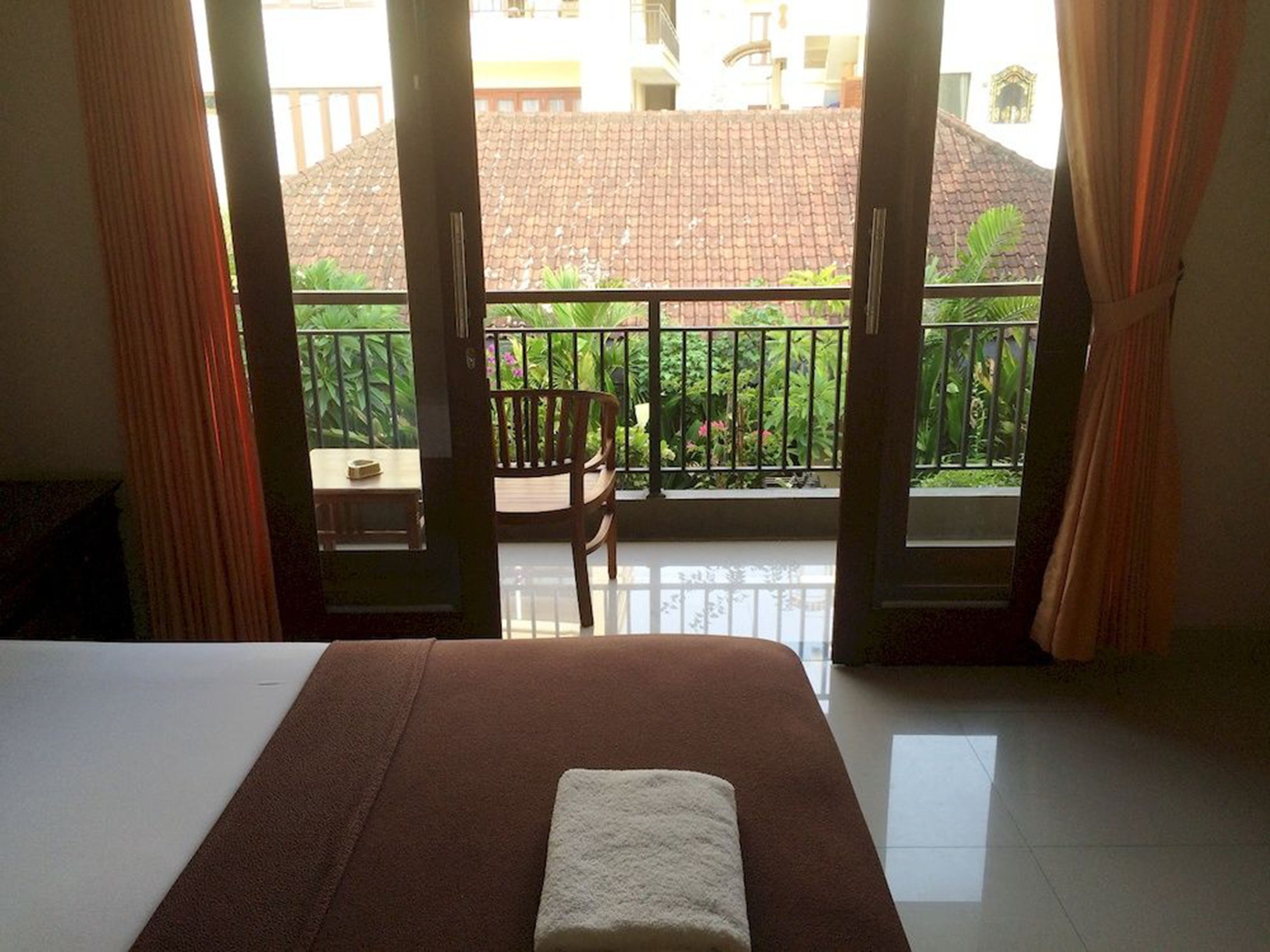Une chambre d’hôtel avec un lit, une petite table et une chaise. La chambre dispose d’un balcon avec vue sur les bâtiments et la verdure.