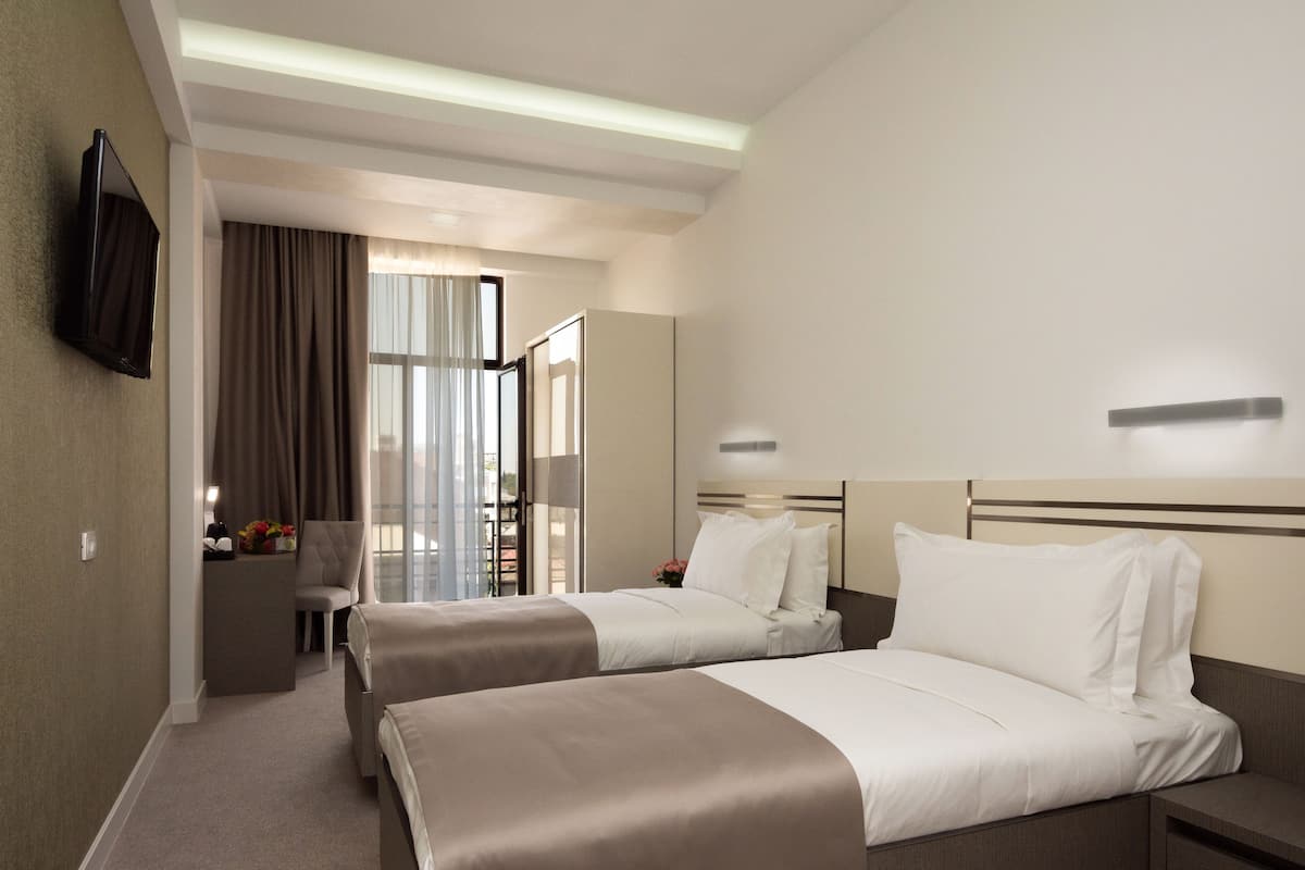 A hotel room with two beds, a desk, a flat-screen TV, and a large window with curtains.
