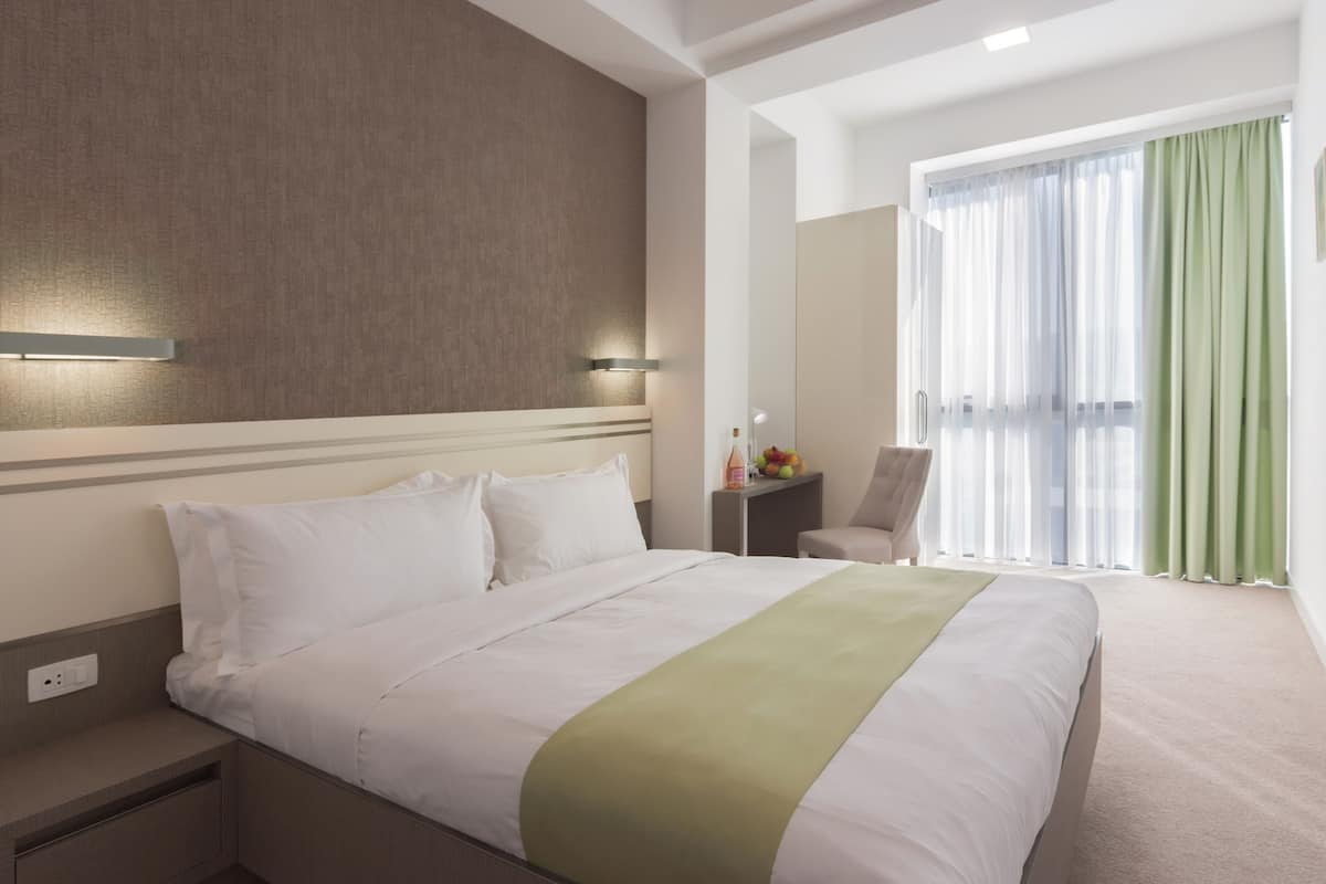 A hotel room with a large bed, a bedside table, a chair, a nightstand, and a window with curtains.