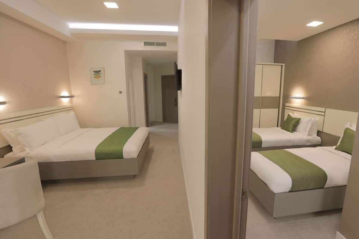 A hotel room with two beds, each with white bedding and a green accent. There is a small desk with a chair, a mirror, and a door leading to another room.