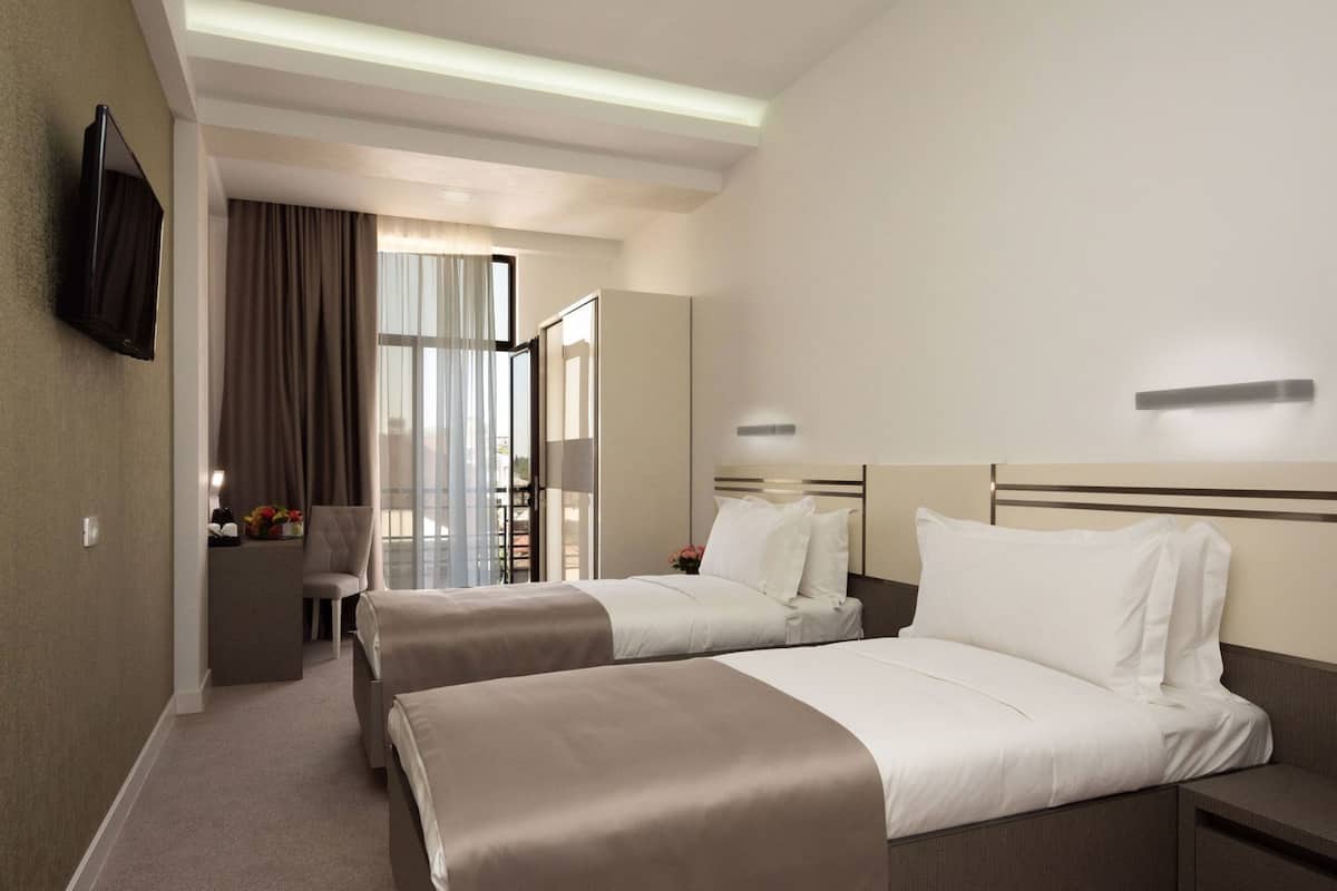 A hotel room with two beds, a desk, a TV, and a large window with curtains.