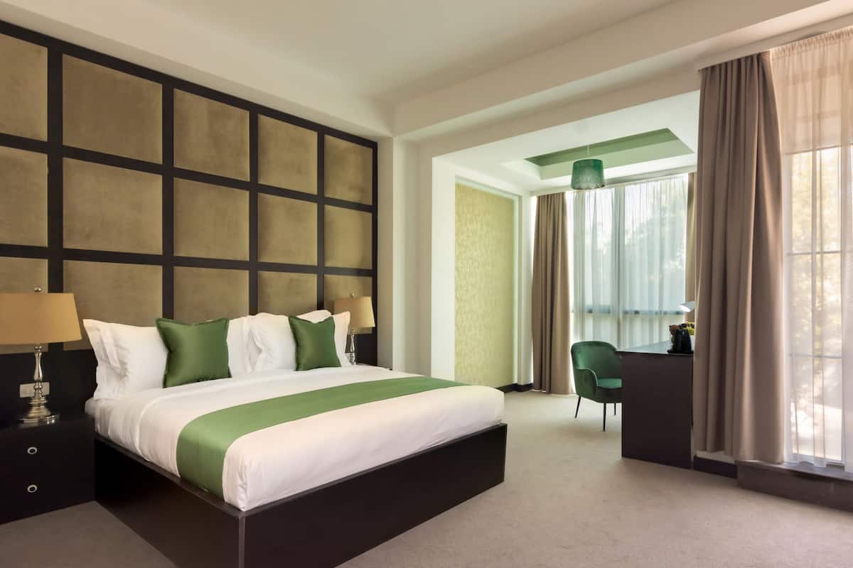 A hotel room with a large bed, a desk, and a chair. There is a window with curtains and a view of the outside.