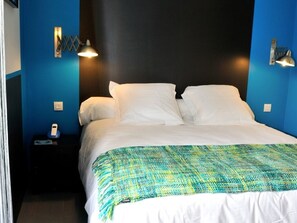 Una cama con ropa de cama blanca y una manta decorativa de color verde y amarillo. Hay dos lámparas de pared a cada lado de la cama.