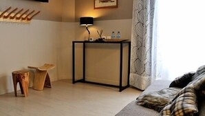Een hotelkamer met een houten vloer, een bank met geruite kussens, een klein houten krukje, een tafel met zwarte lijst en een lamp, een schilderij aan de muur, en een raam met gordijnen met een patroon.