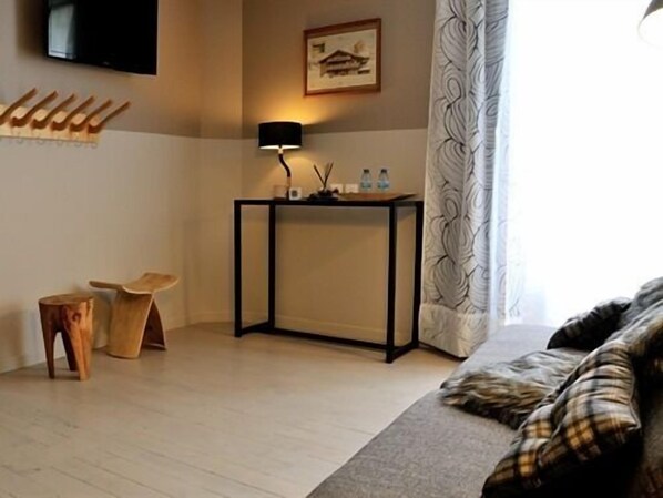 Une chambre d’hôtel avec un plancher en bois, un canapé avec des coussins à carreaux, un petit tabouret en bois, une table à cadre noir avec une lampe, un tableau au mur et une fenêtre avec des rideaux à motifs.