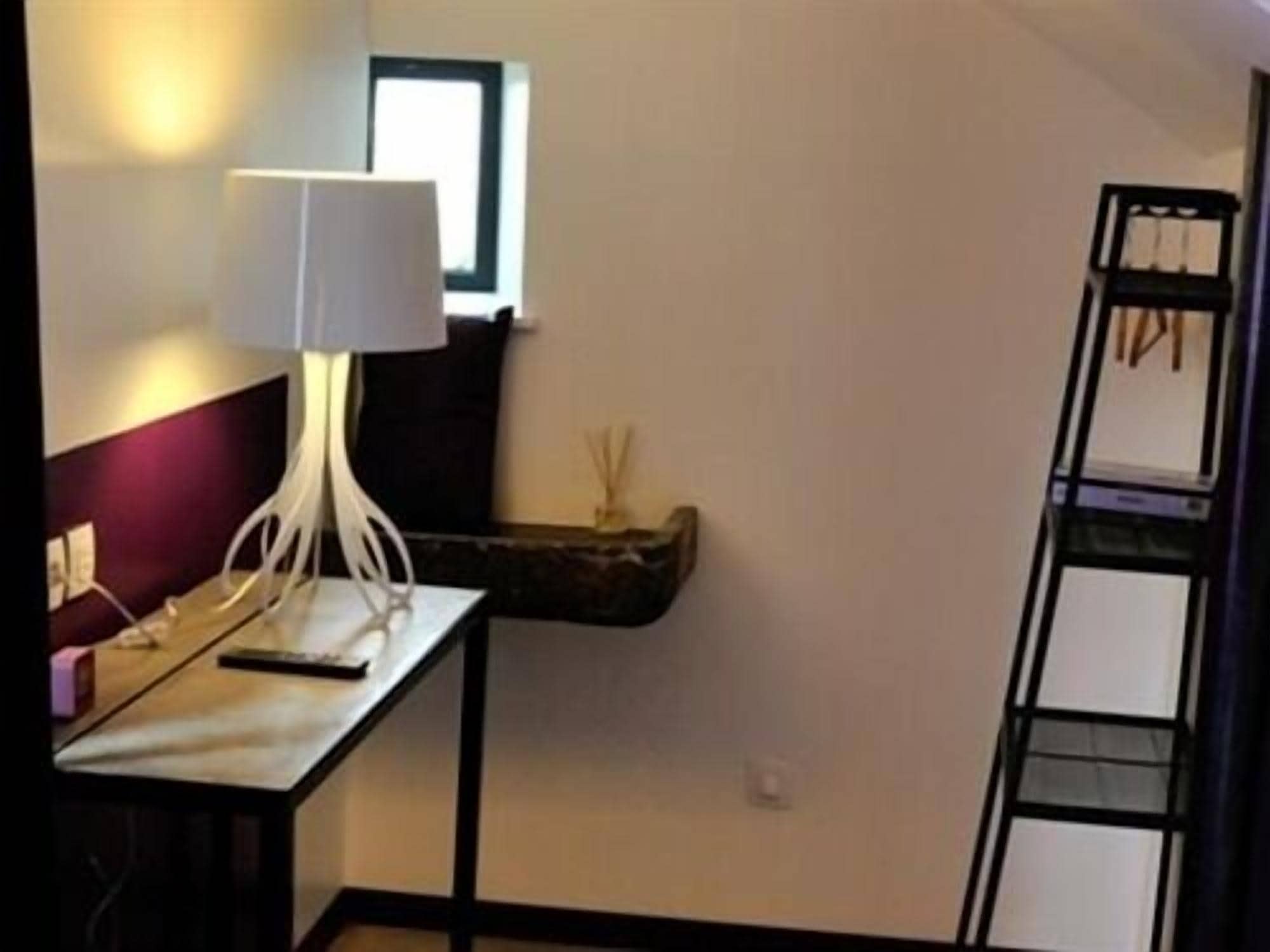 Una habitación con escritorio, lámpara, un pequeño estante con adornos y una escalera apoyada en la pared.