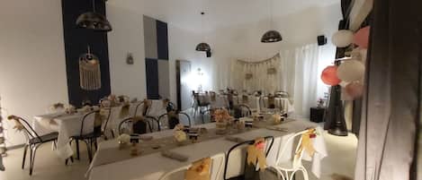 Un comedor con varias mesas dispuestas para una comida, sillas alrededor y elementos decorativos como luces colgantes y adornos en las paredes.