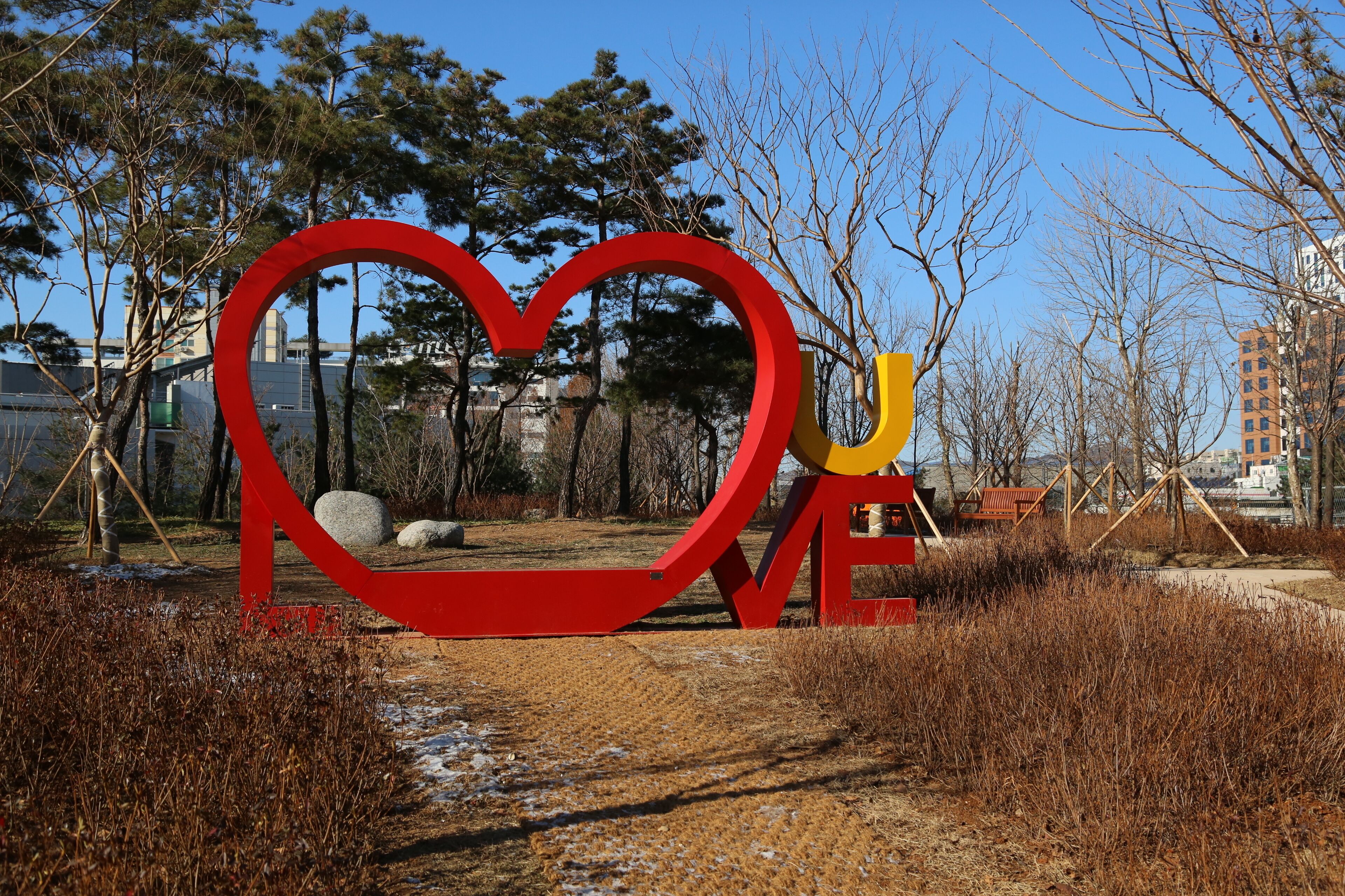 A heart-shaped sculpture in a park with trees and a building in the background.