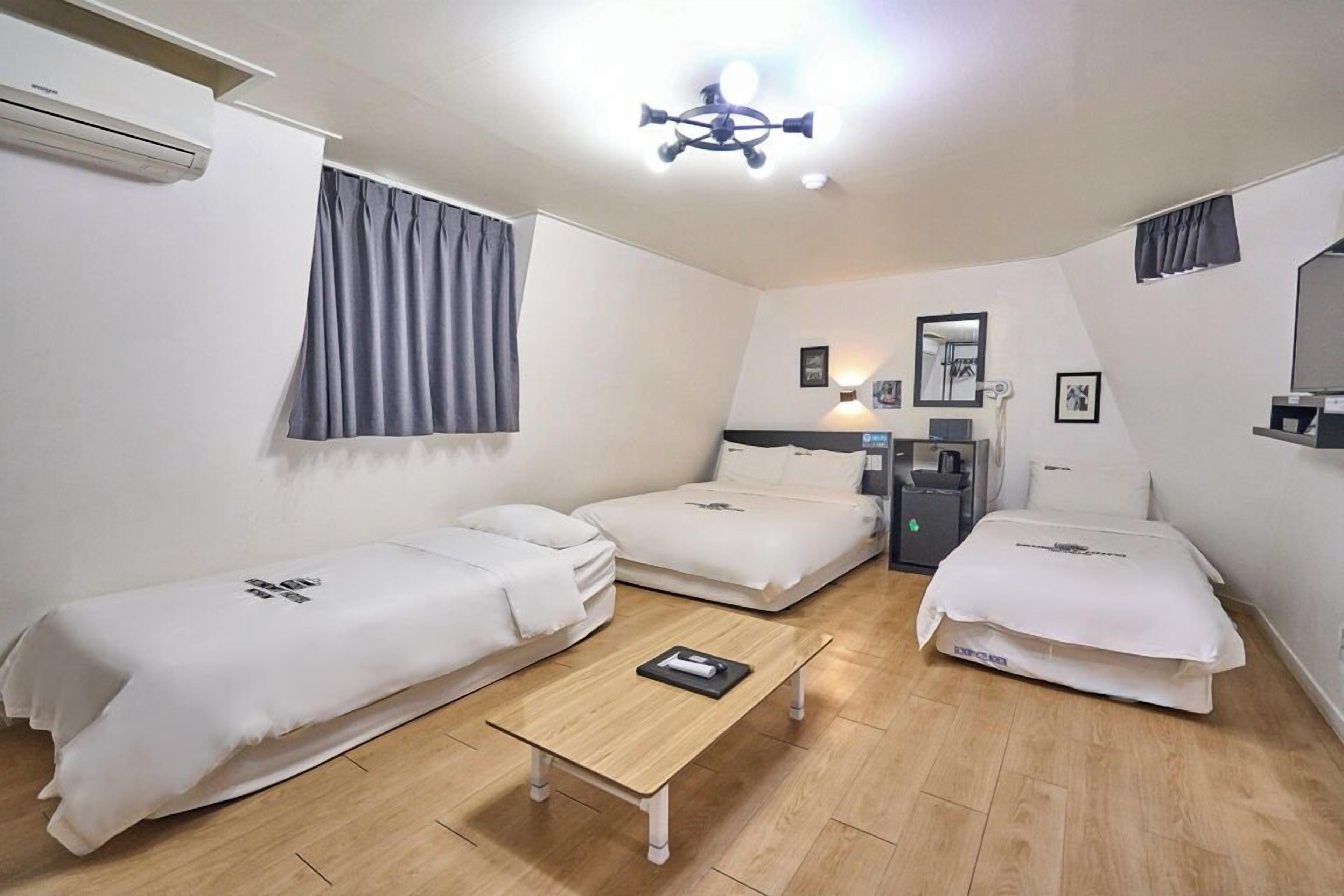 A compact hotel room with two single beds, a small desk, a chair, and a bedside table.