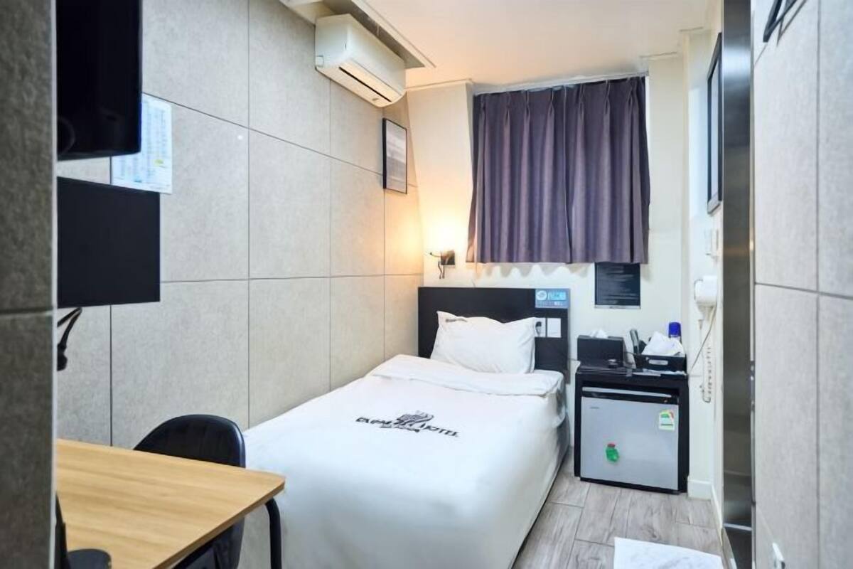 Une chambre d’hôtel comprenant un lit, un bureau, une chaise, une petite table, un téléviseur, un climatiseur, une fenêtre avec des rideaux et une table de chevet avec une lampe.