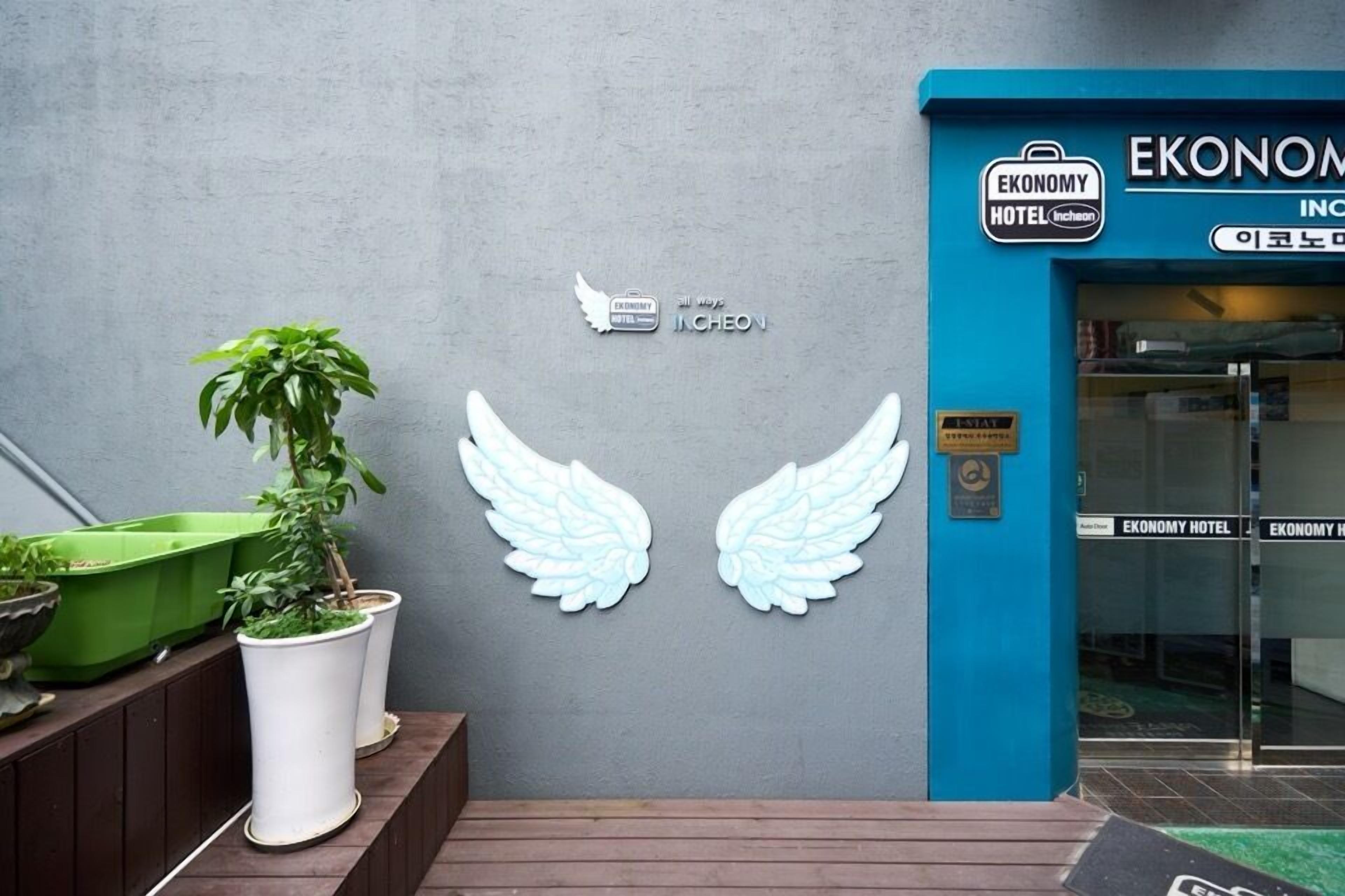 Een hotel ingang met een blauwe gevel, een bord met 'Economy Hotel' en een decoratieve set witte engel vleugels.
