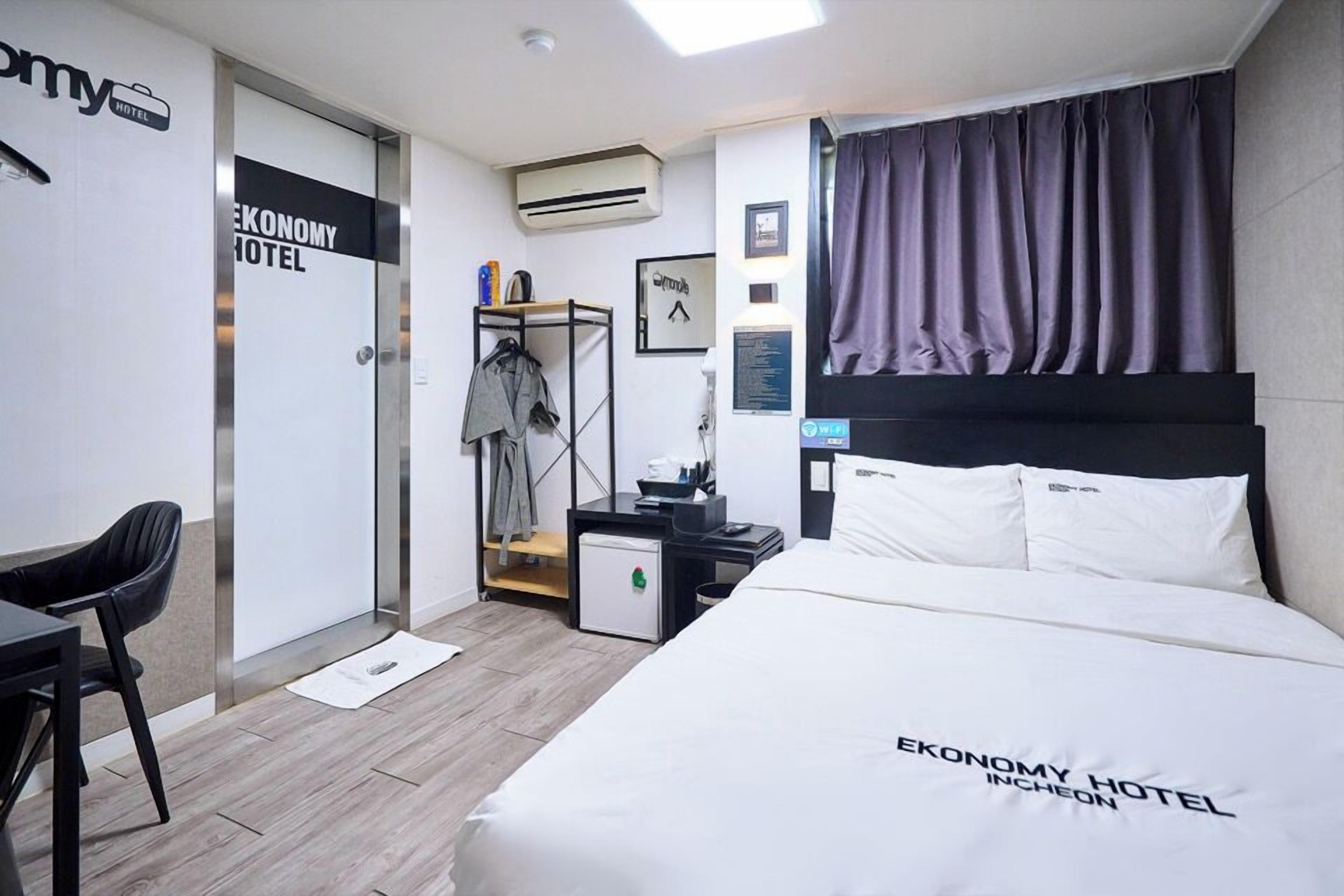 A hotel room with a bed, a desk, a chair, a wardrobe, and a small table with a lamp.