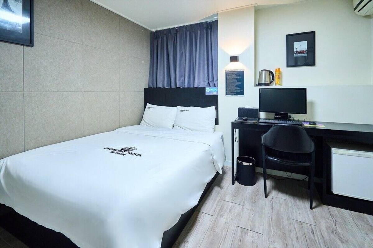 Une chambre d’hôtel comprenant un lit, un bureau avec un ordinateur, une chaise, une poubelle et un tableau accroché au mur.
