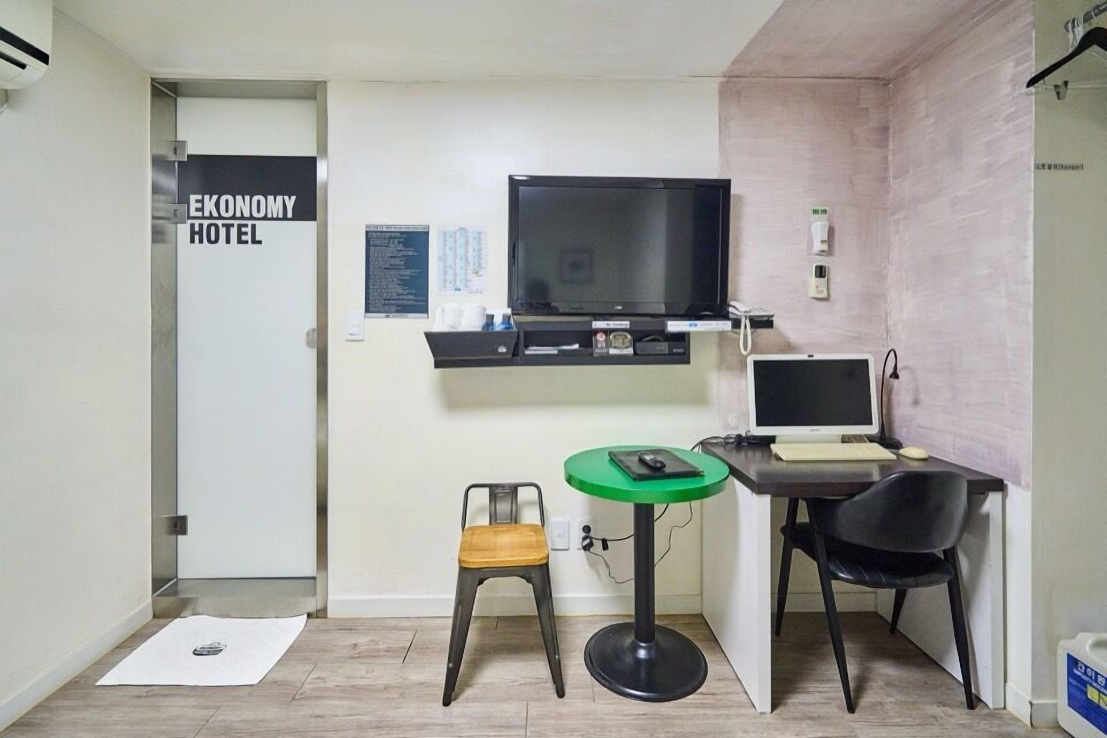 A small room with a television, a desk with a computer, a chair, and a small stool. There is a sign on the door that reads 'Economy Hotel.'