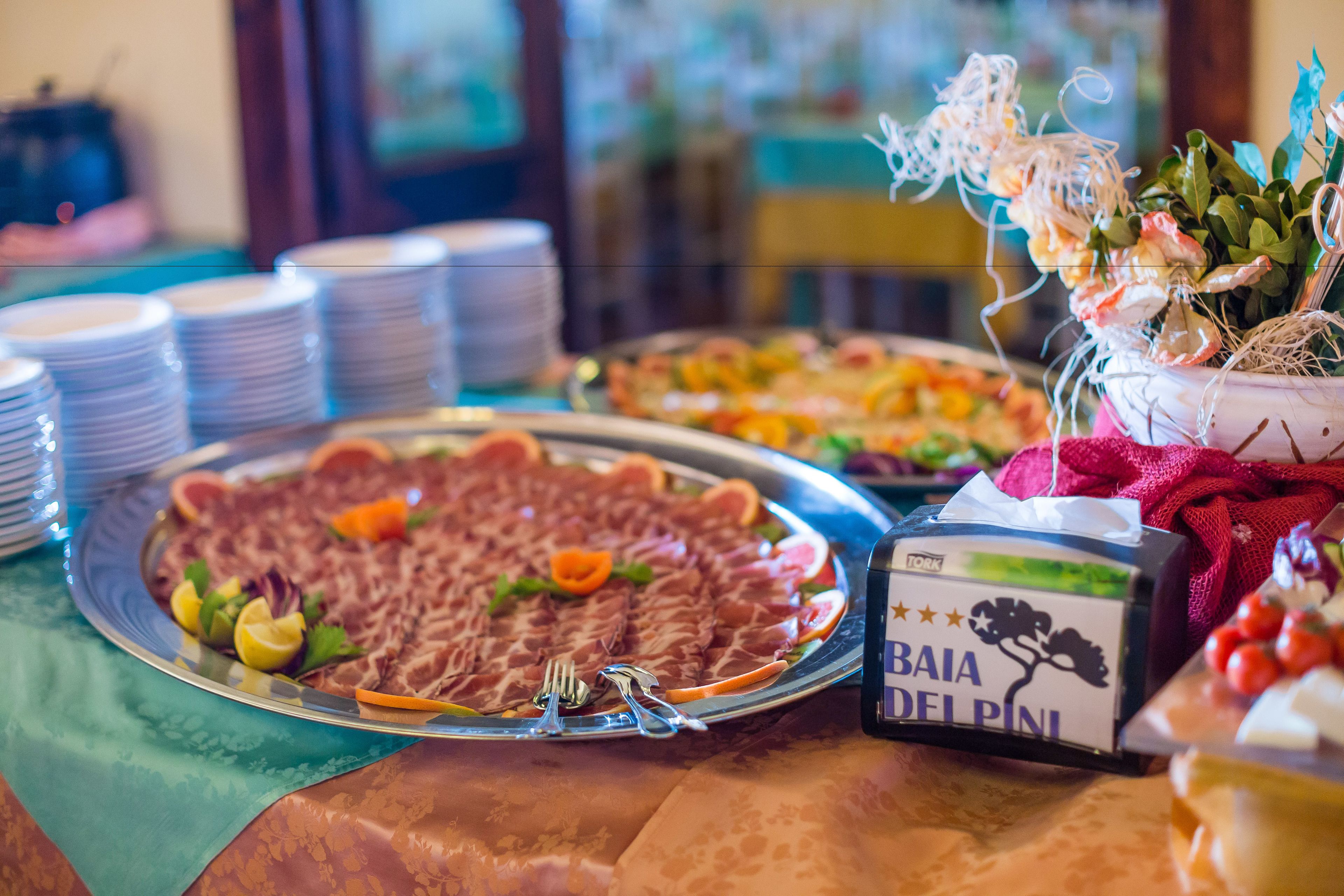 A buffet table with various dishes, including a large platter of sliced meats, a box labeled "BAIA DEL PINI," and a floral arrangement.