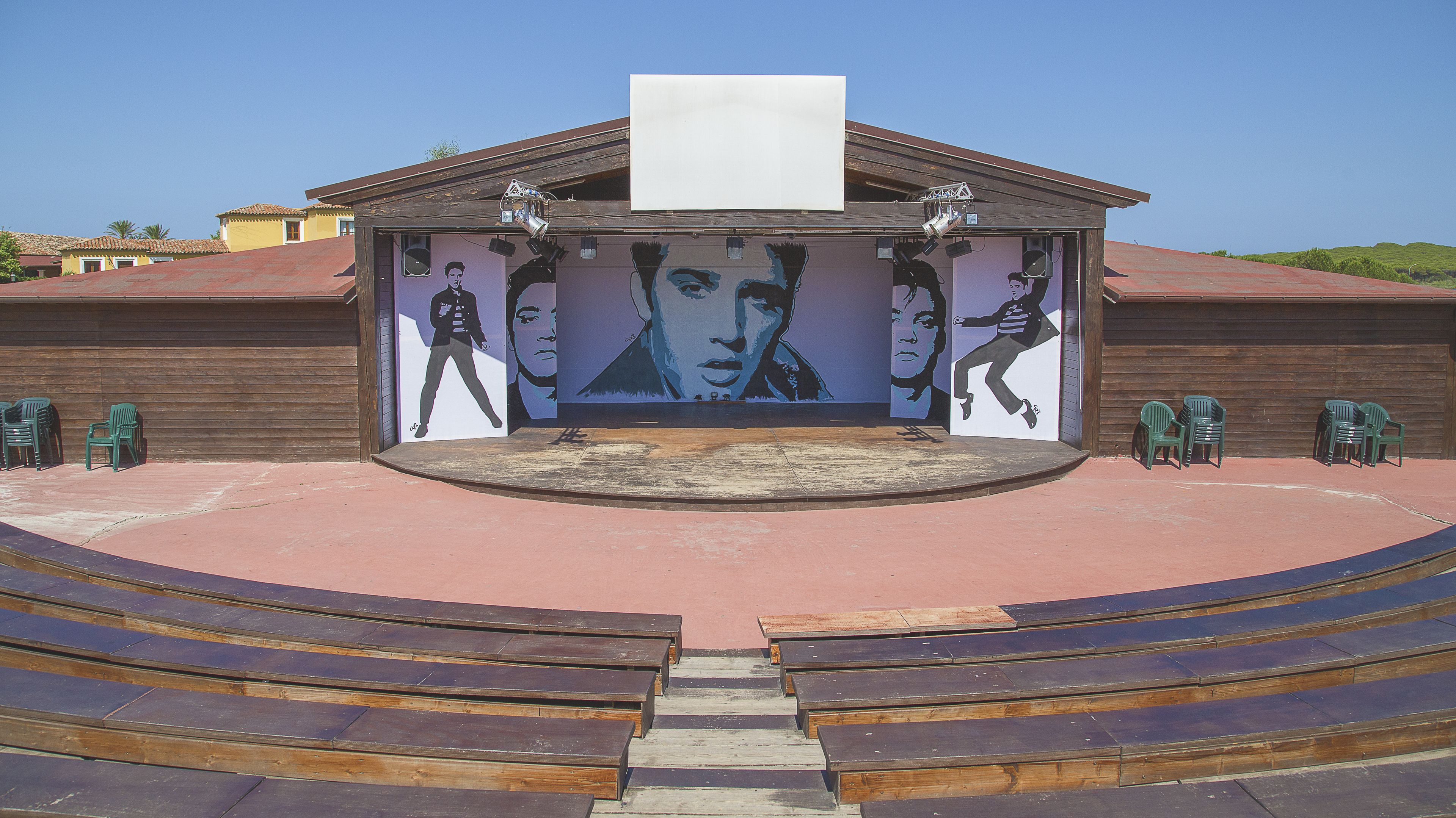 An outdoor amphitheater with a stage featuring a large mural, surrounded by green plastic chairs.