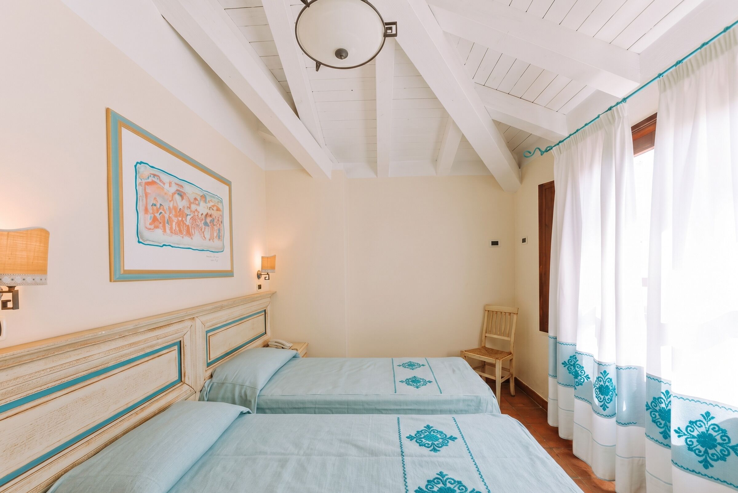 A room with two beds, a framed picture on the wall, a lamp, and a window with curtains.