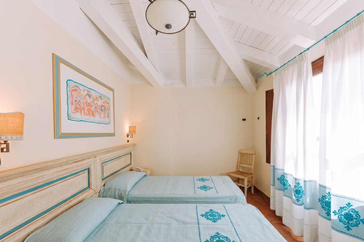 A room with two beds, a framed picture on the wall, a lamp, and a window with curtains.