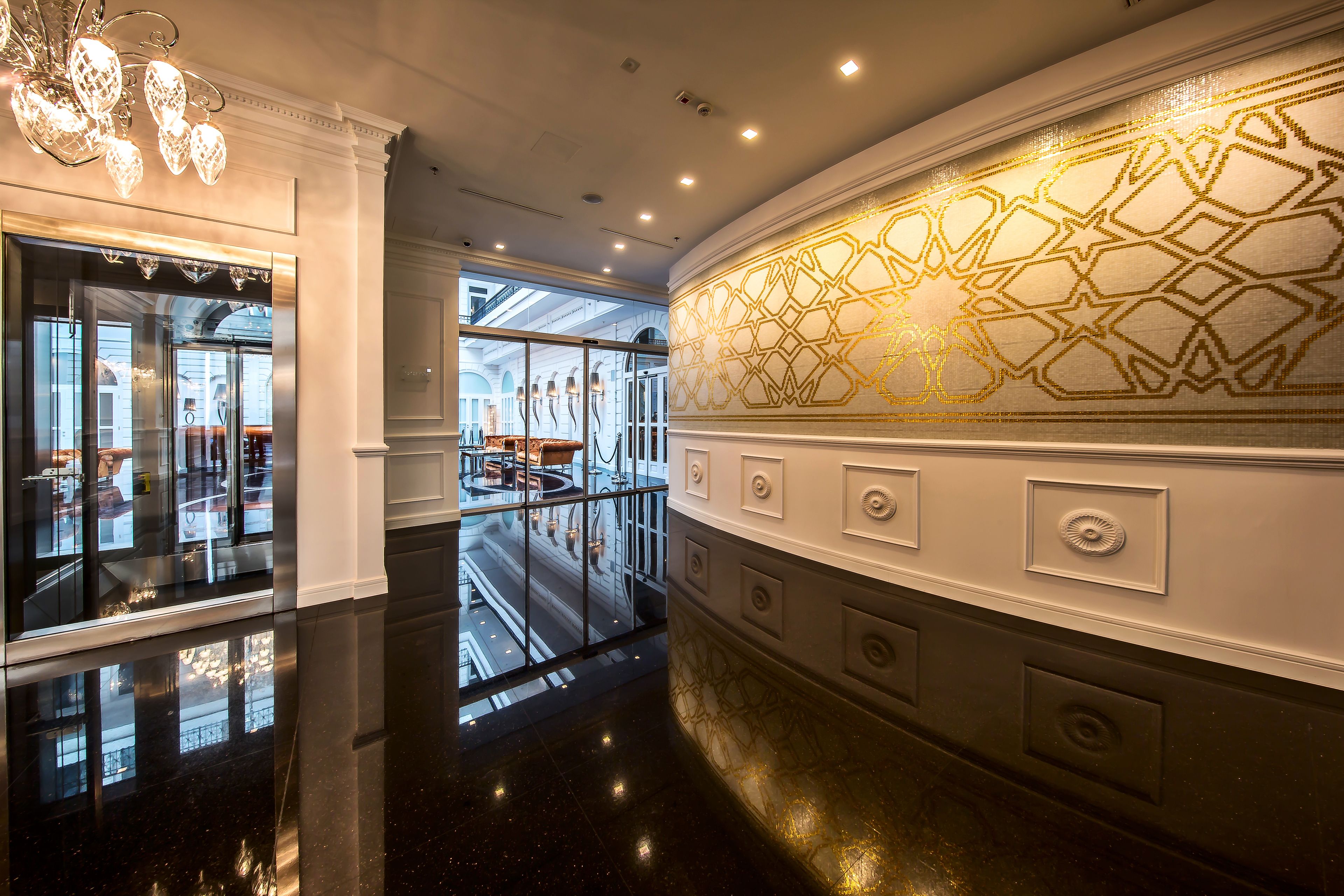 A polished lobby with reflective floors, a glass elevator, and a decorative wall with geometric patterns.
