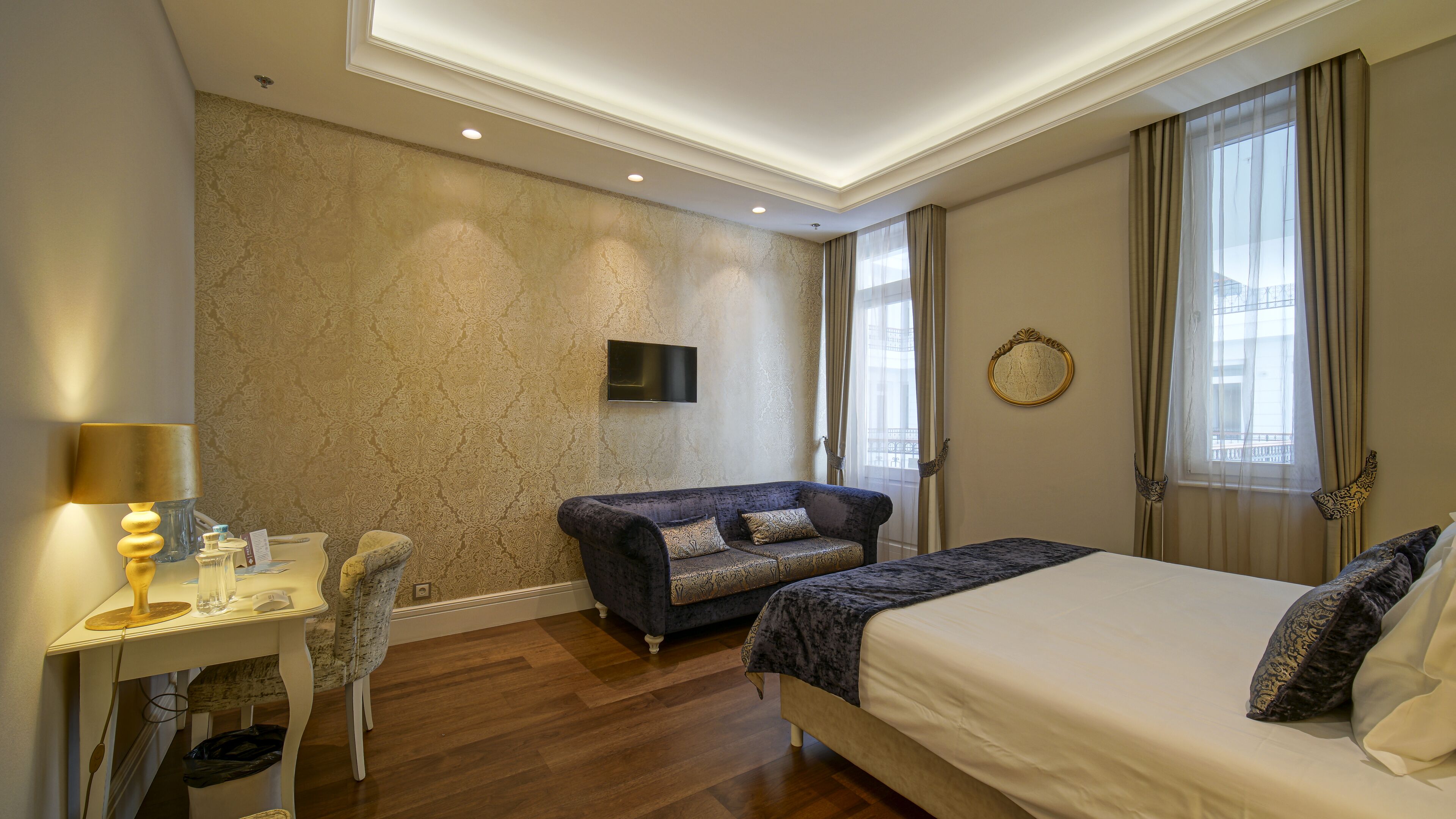 A hotel room with a bed, a sofa, a desk with a lamp, a TV, and a window with curtains.