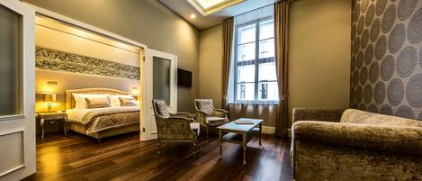 Habitación de hotel con cama, dos sillones, una mesita, un escritorio con lámpara y un amplio ventanal con cortinas.