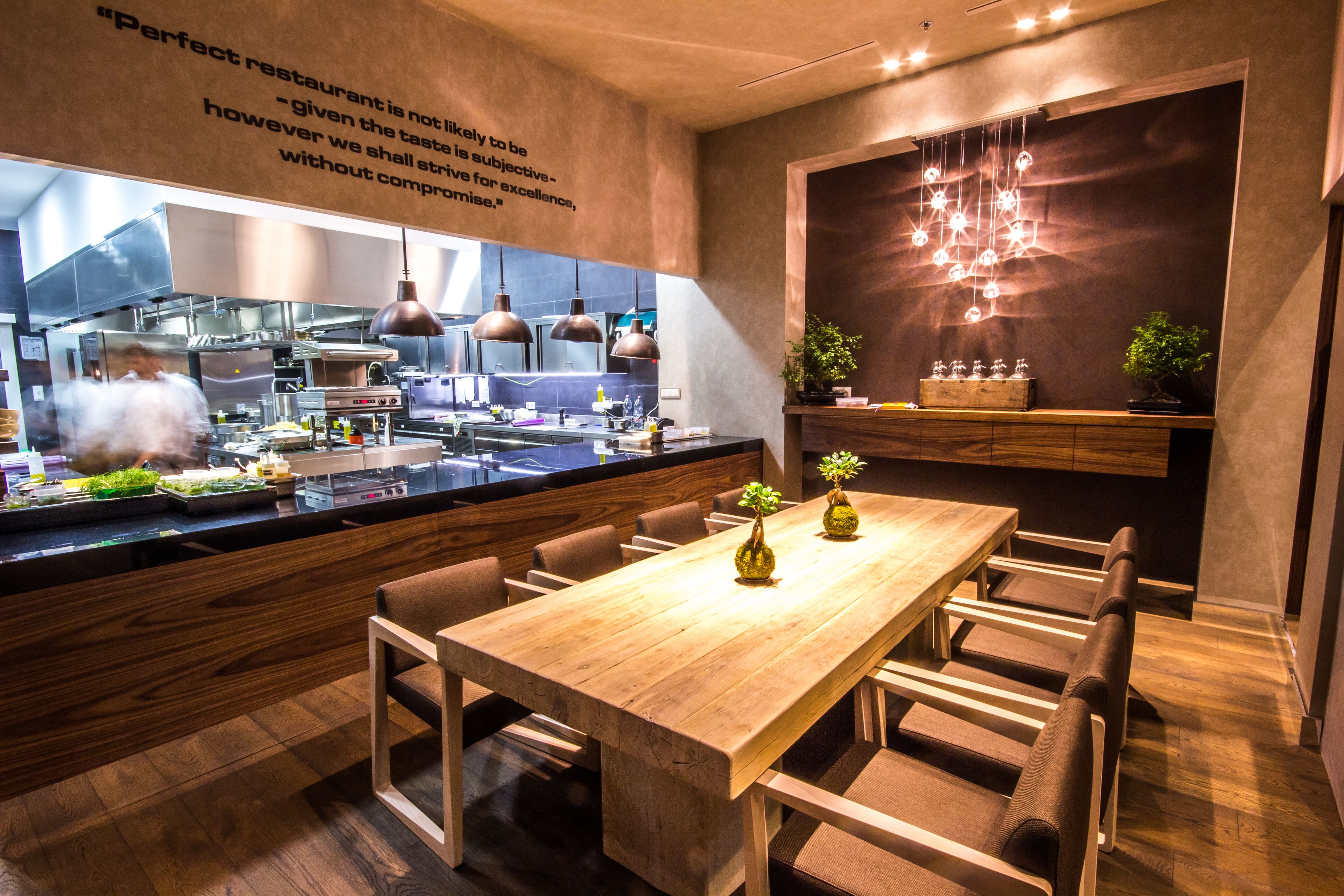 A modern restaurant interior with a long wooden table, comfortable seating, a kitchen in the background, and decorative lighting.