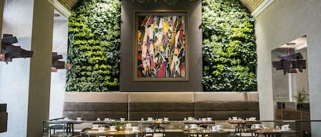 Un restaurante con una decoración moderna, que presenta una pared verde con plantas, un cuadro abstracto grande y mesas y sillas ordenadas.