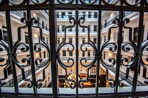 A view through ornate iron gates of a multi-story building with balconies and a central chandelier.