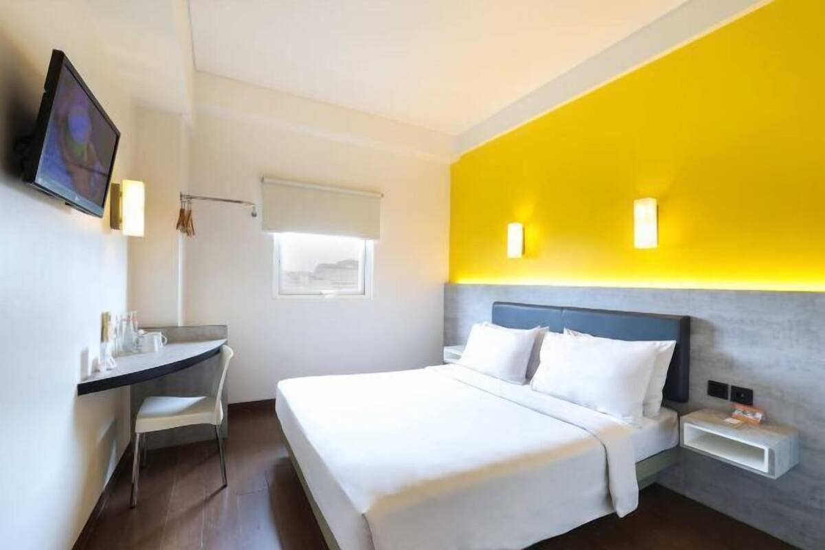 Une chambre d’hôtel équipée d’un lit, d’une table de chevet, d’une chaise, d’une télévision et d’une fenêtre avec des stores.