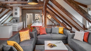 A loft-style living room with exposed wooden beams, grey sofas with patterned cushions, a white coffee table, and a kitchen area in the background. - 4 Arts Apartments by Adrez Living (Prague)