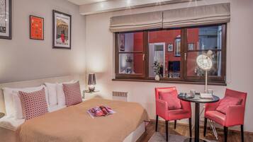 Habitación de hotel con cama, mesita de noche con lámpara, una silla roja, una pequeña mesa redonda con una revista y una ventana que da a otra habitación.
