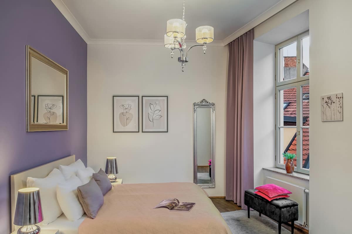 Une chambre à coucher comprenant un lit, une lampe de chevet, un miroir, une œuvre d’art et une fenêtre donnant sur un paysage.