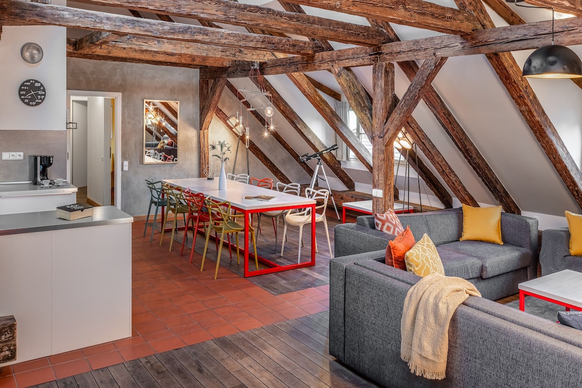 Une pièce de style « loft » avec des poutres en bois apparentes, un coin repas doté d’une table à cadre rouge et de chaises colorées, ainsi qu’un canapé gris agrémenté de coussins jaunes.