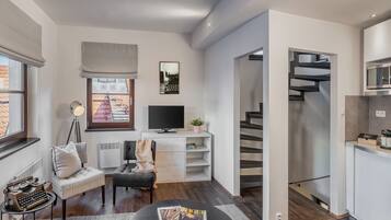 A modern living room with a flat-screen TV, a comfortable sofa, a small round table with a magazine, a floor lamp, and a staircase leading to the kitchen.