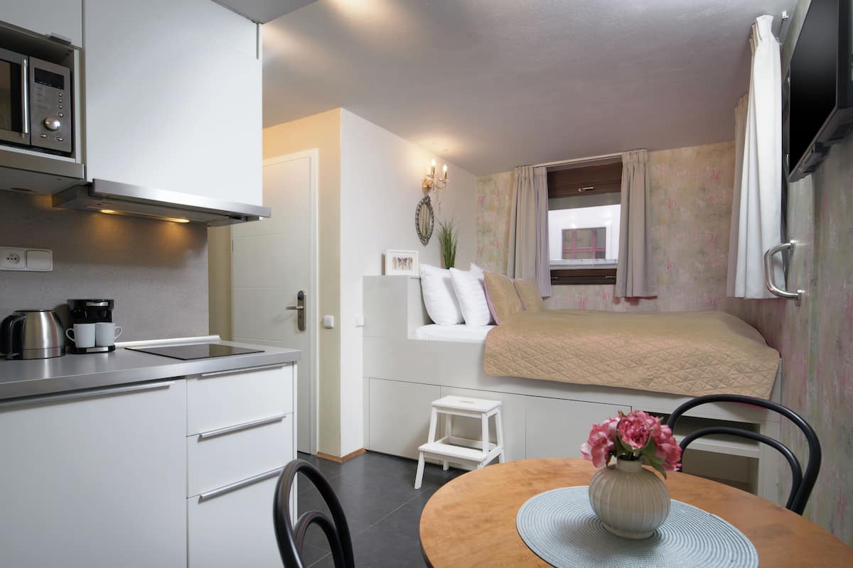Une chambre d’hôtel compacte comprenant un coin cuisine, un lit près d’une fenêtre, une petite table avec un vase et une chaise.