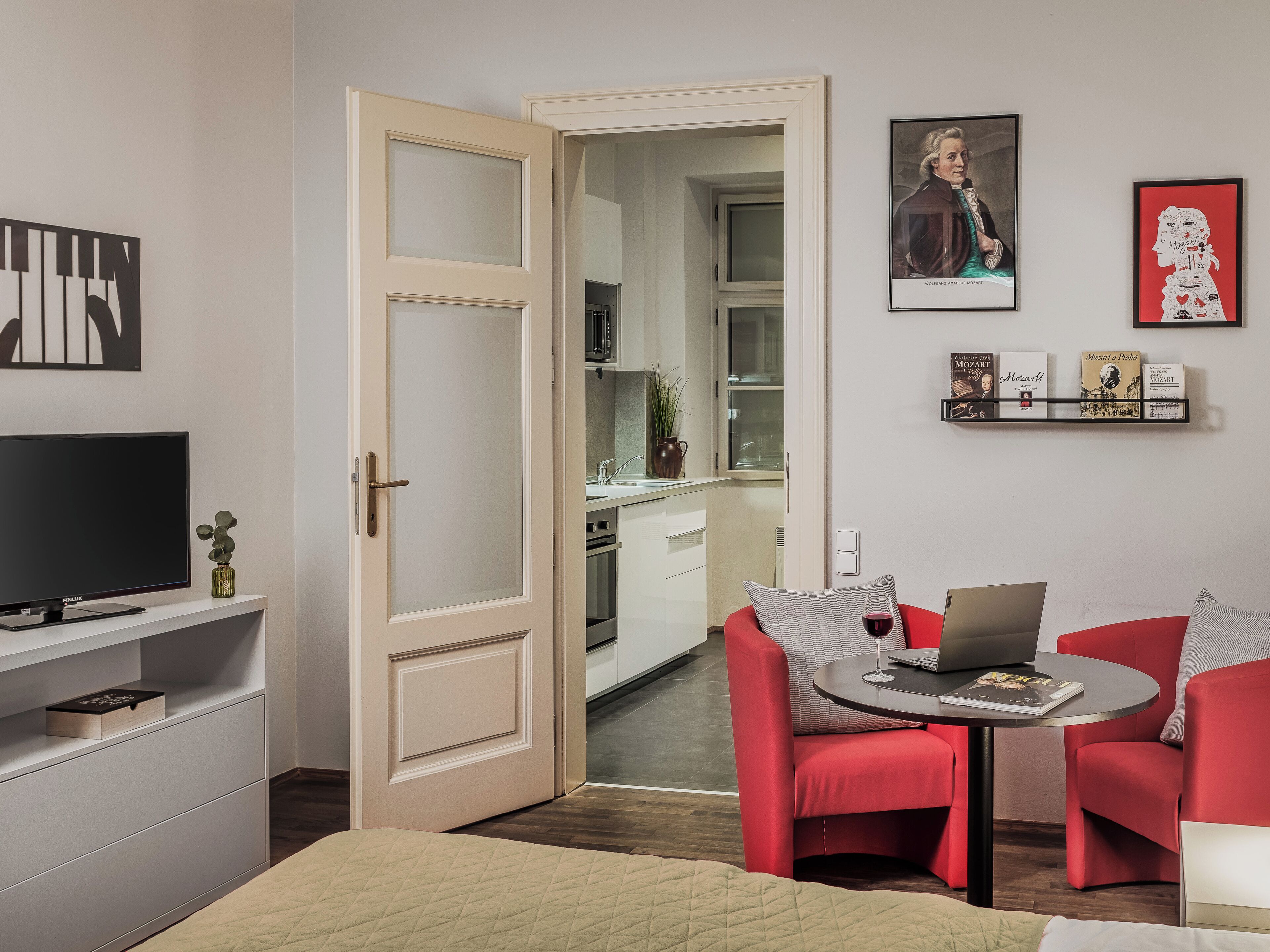 A modern living room with a TV, red chairs, a small table with a laptop and wine, and a kitchen visible through an open door.