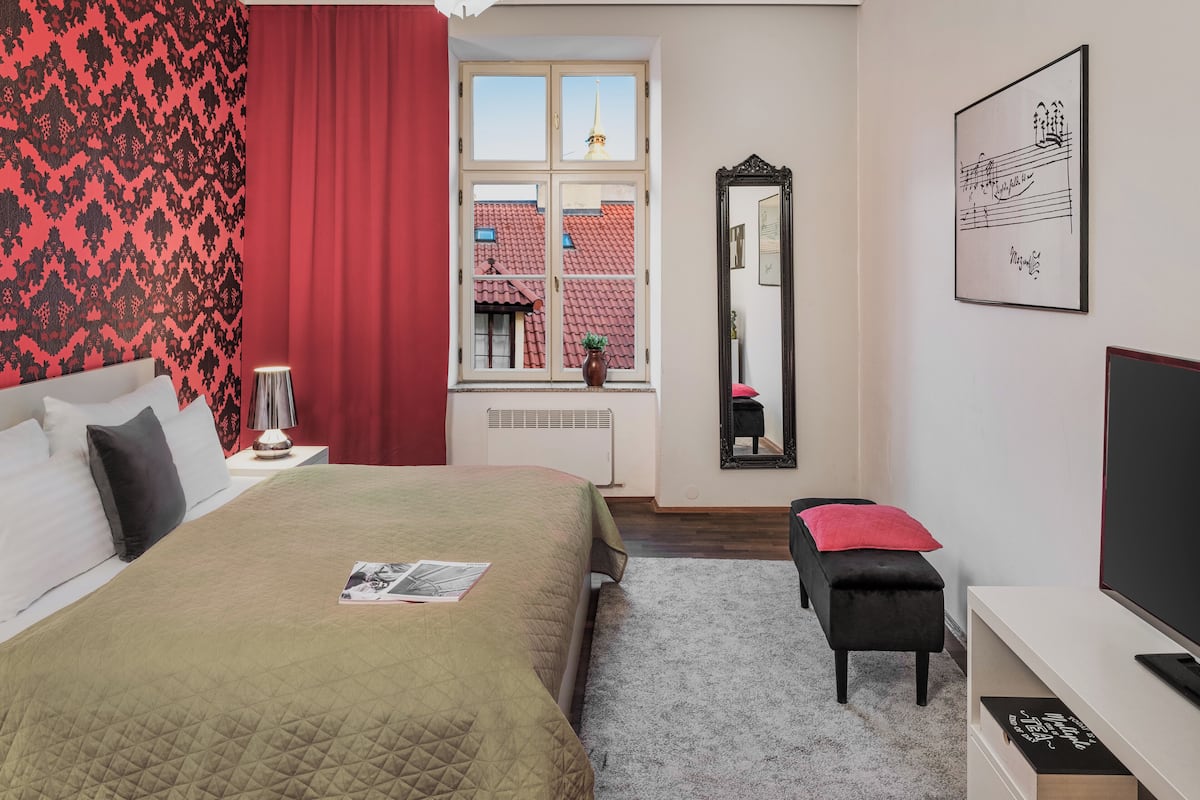 Une chambre d’hôtel avec un lit, une table de chevet avec une lampe, une fenêtre donnant sur des immeubles, un miroir et un tableau encadré au mur.