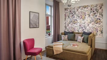 Een gezellige kamer met een bank, een rode stoel, een raam met gordijnen, bloemenbehang en een kroonluchter.