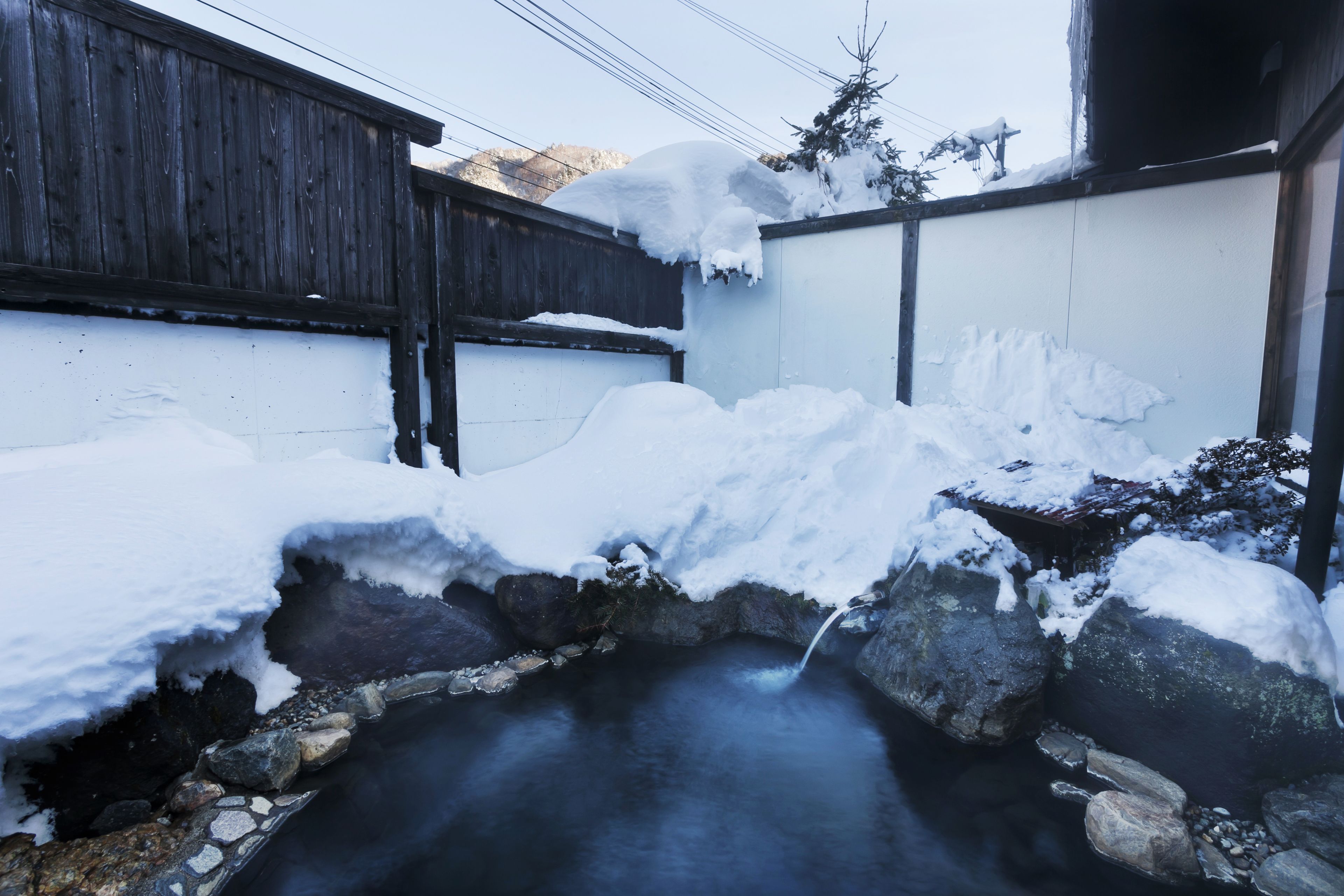 A stone hot spring surrounded by snow, with a wooden fence and a building in the background.